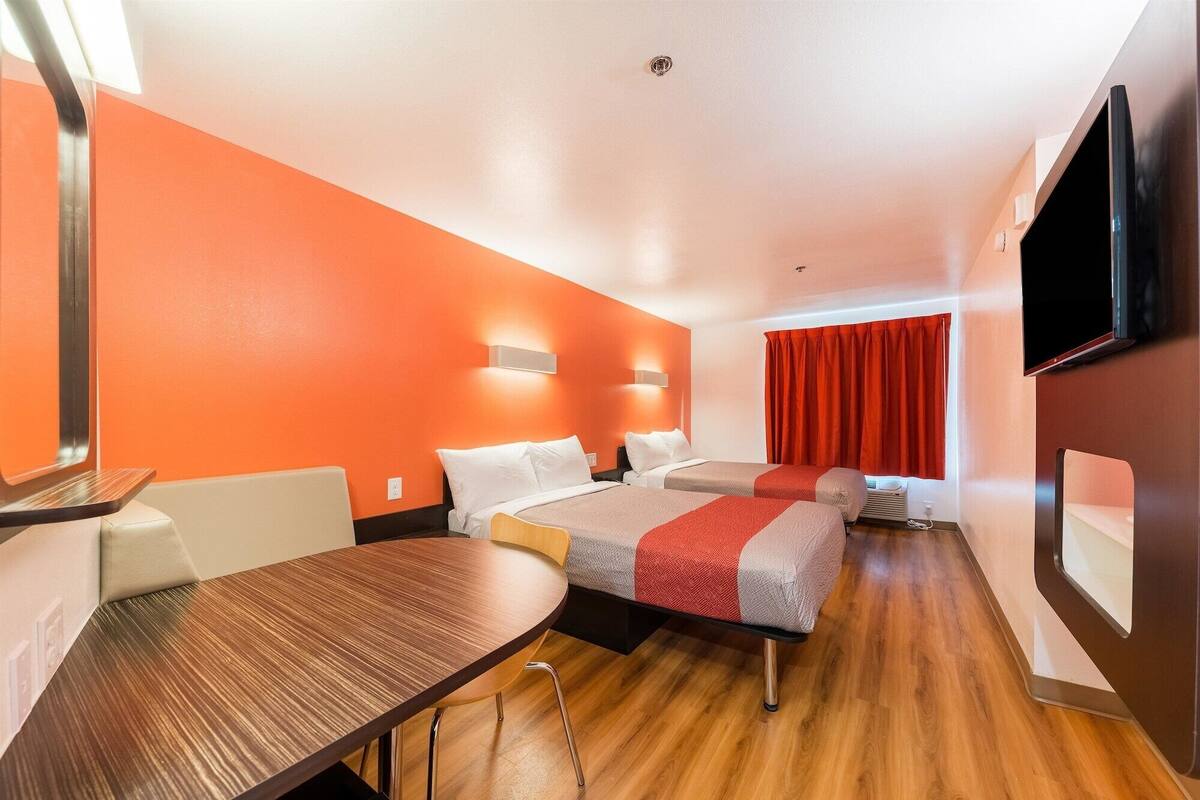 A hotel room with two beds, a wooden table, a TV, and a window with red curtains.