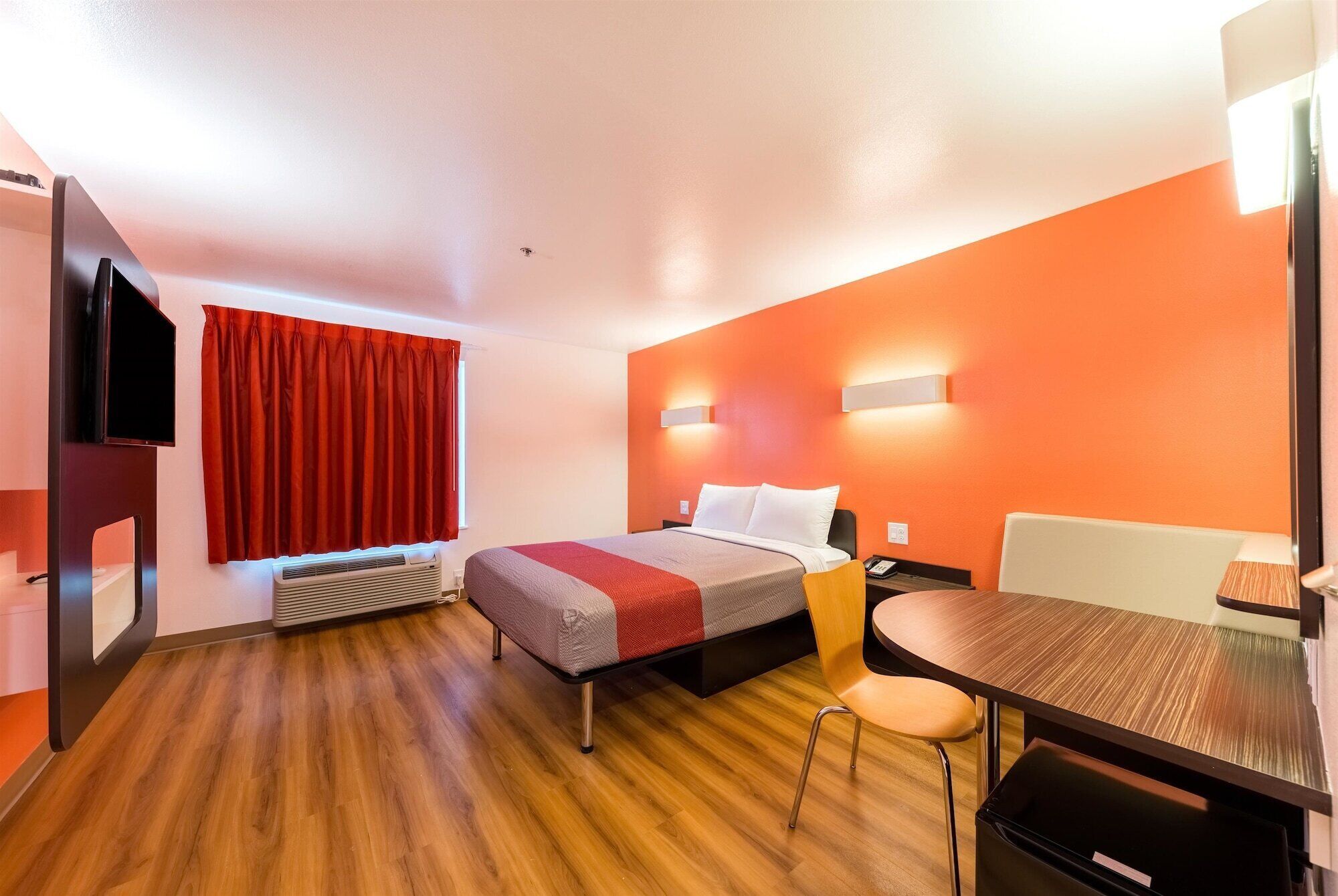 Una habitación de hotel con cama, escritorio, silla, televisor y ventana con cortinas rojas.