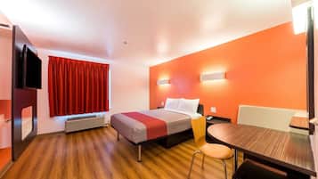 Una habitación de hotel con cama, escritorio, silla, televisor y ventana con cortinas rojas.