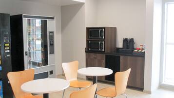 Une petite cuisine équipée d’un four à micro-ondes, d’une machine à café et d’un réfrigérateur. On y trouve deux tables rondes blanches et quatre chaises orange.