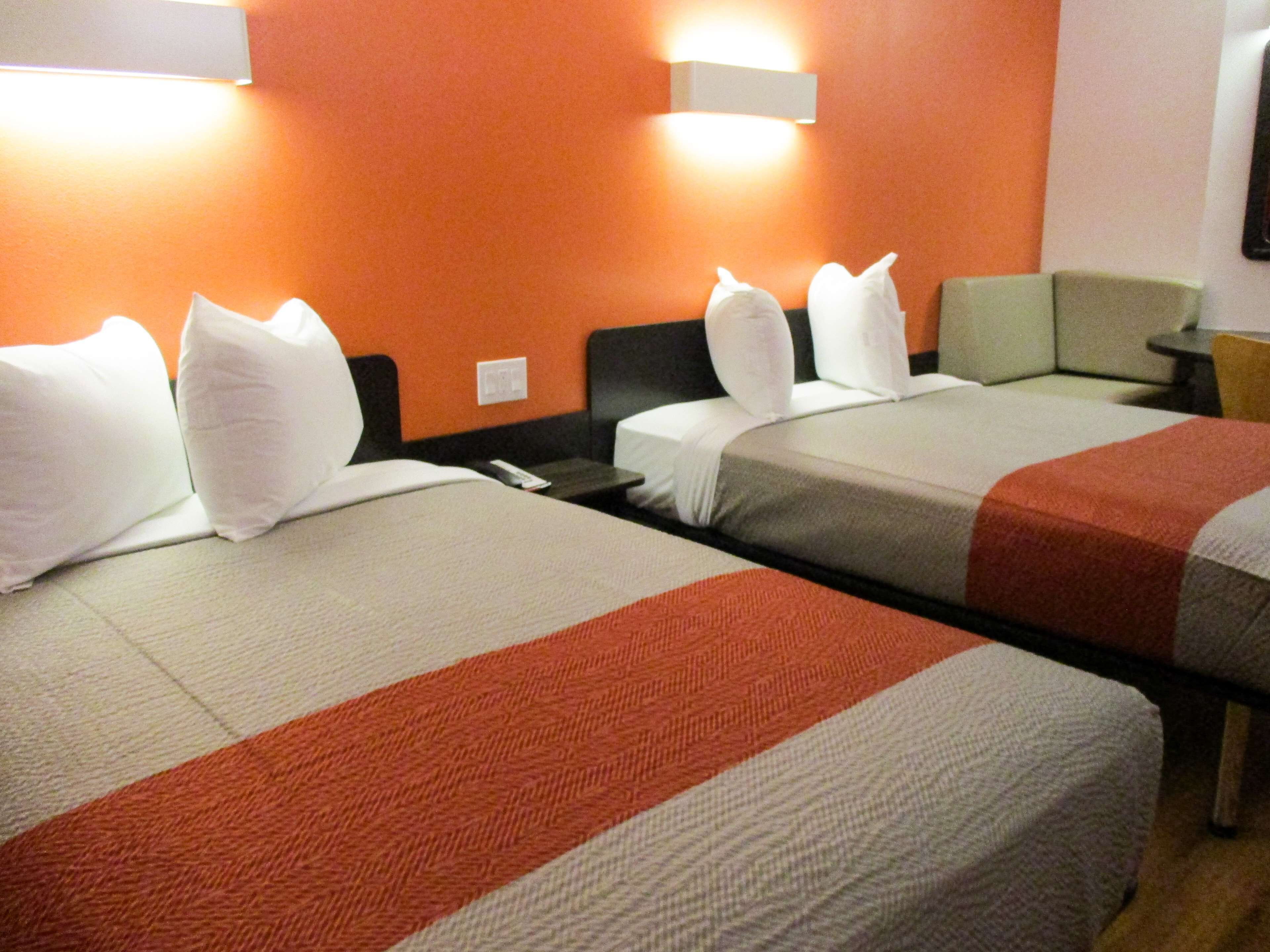 A hotel room with two beds, each with white pillows and a red and grey bedspread. There is a nightstand with a lamp and a small table with a chair.