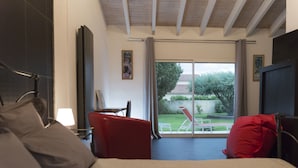Un dormitori amb un llit, una cadira vermella, una taula petita i vista a un jardí per una porta corredissa.