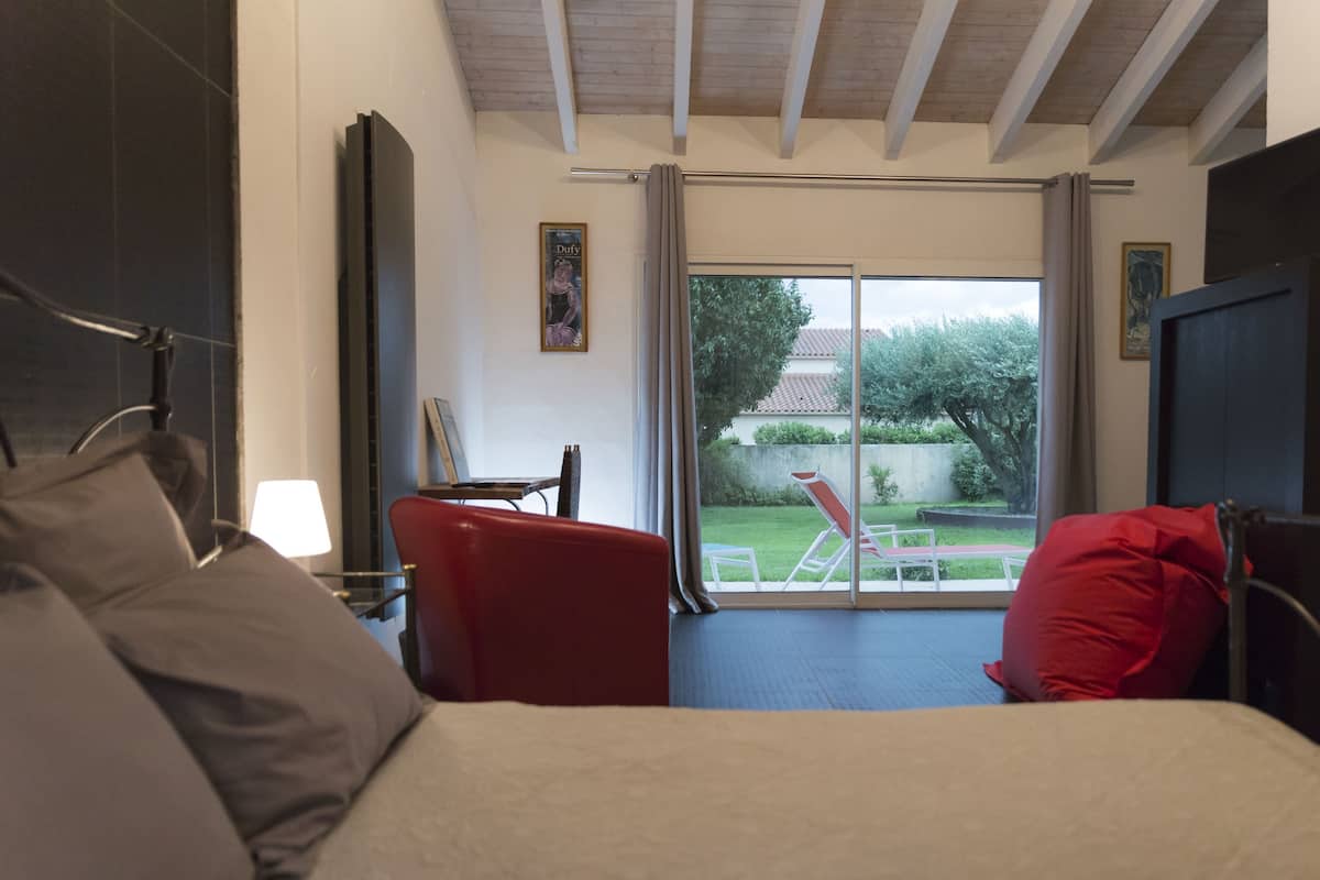 A bedroom with a bed, a red chair, a small table, and a view of a garden through a sliding door.