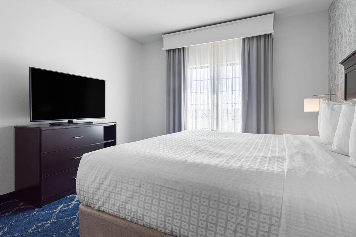 Hotellihuone, jossa on sänky, tummalla lipastolla oleva taulutelevisio, ikkuna, jossa on verhot, ja yöpöydällä oleva lamppu.