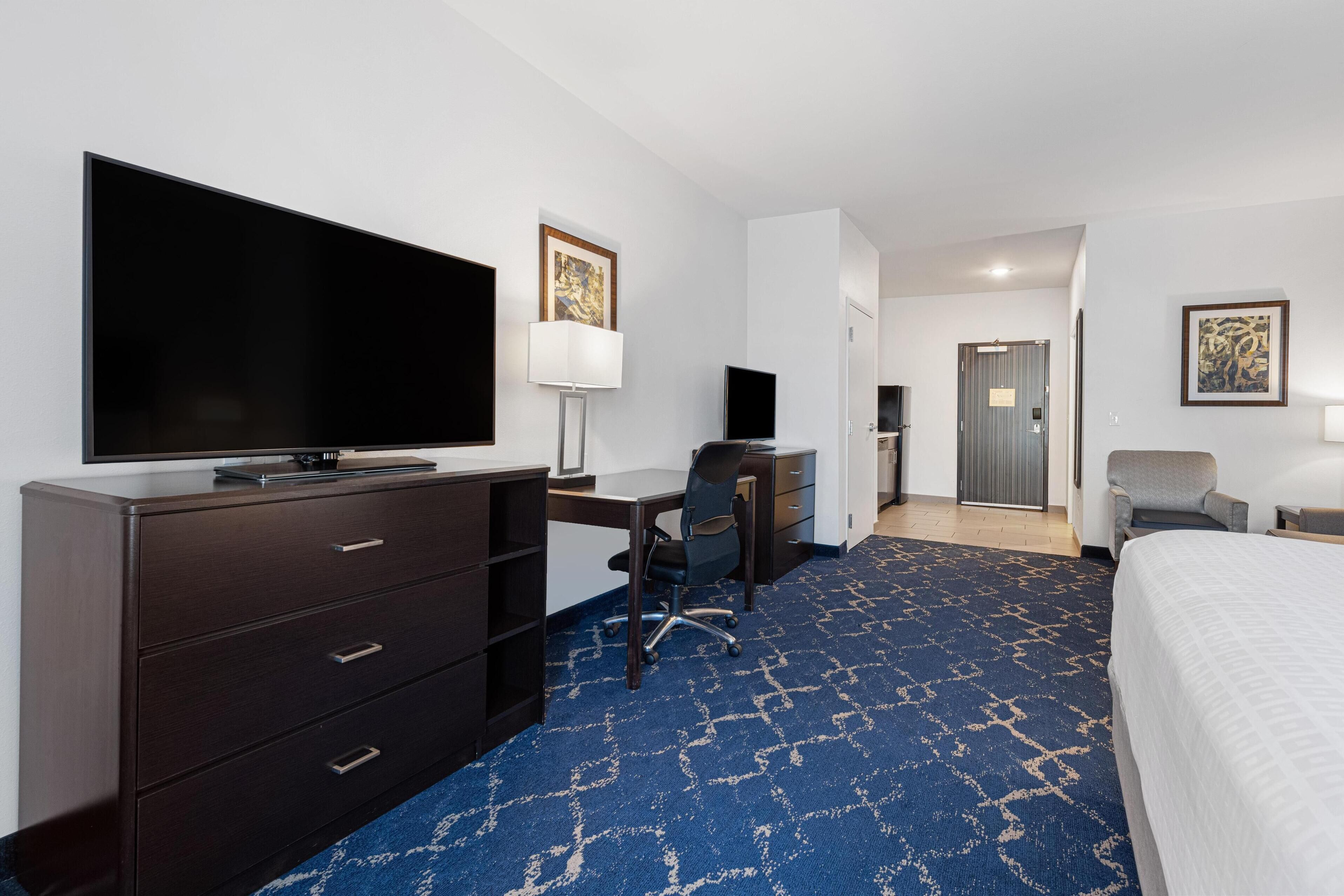 A hotel room with a large flat-screen TV, a desk with a chair, a bed, and a blue patterned carpet.