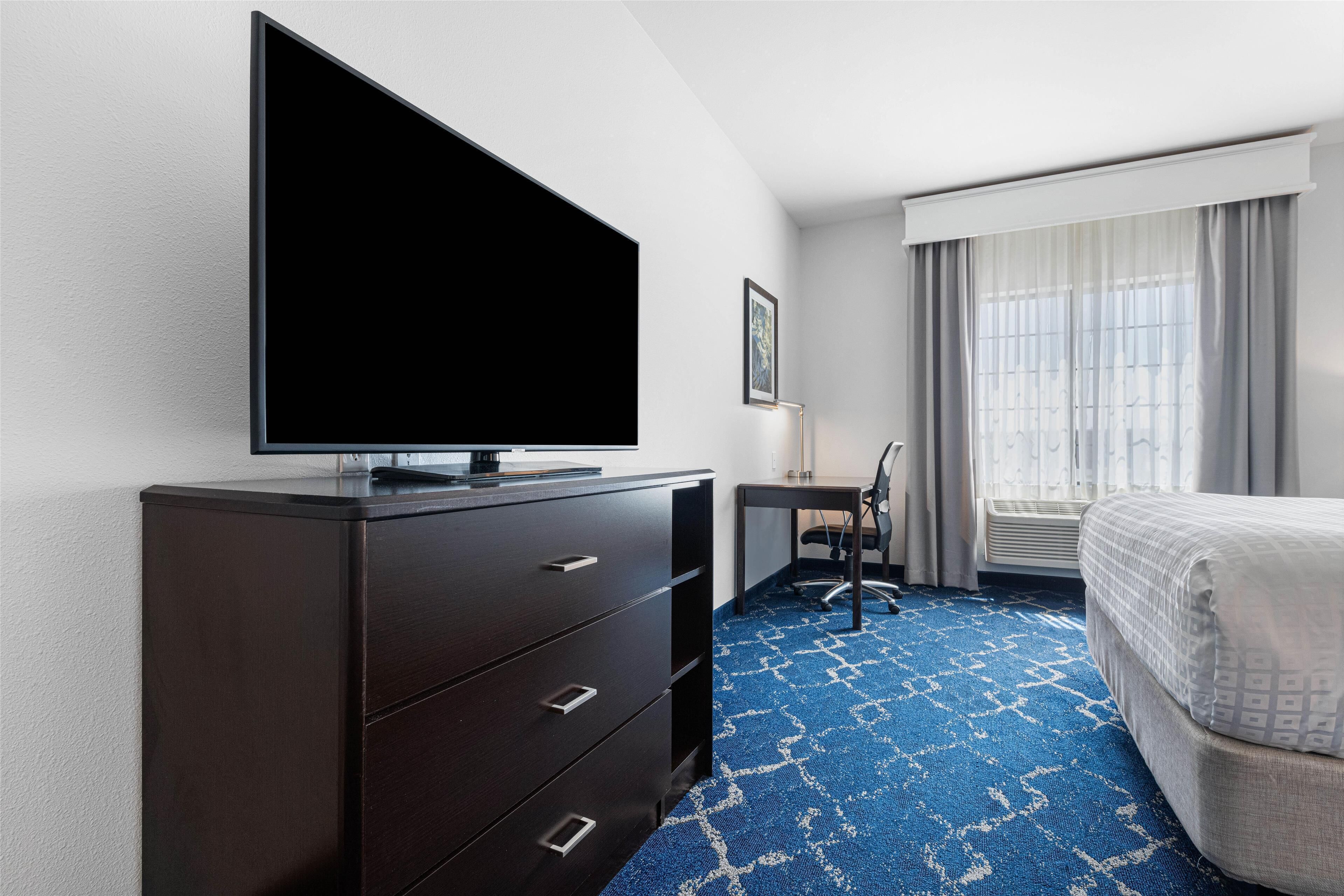 A hotel room with a large flat-screen TV on a dark wooden dresser, a desk with a chair, a bed with a white comforter, and a blue patterned carpet.