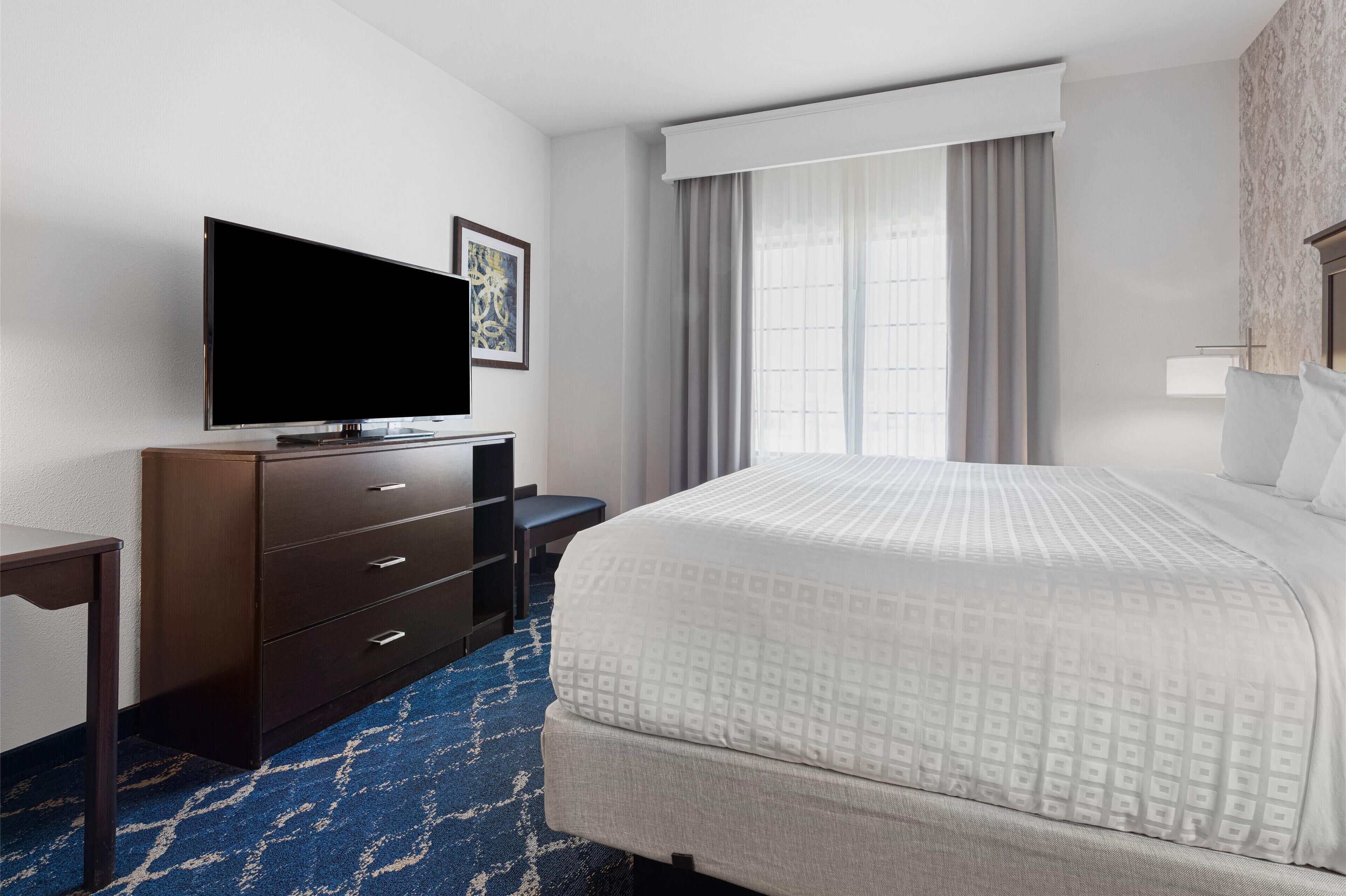 Habitación de hotel con una cama grande, un televisor de pantalla plana sobre un tocador de madera, un escritorio y una ventana con cortinas.