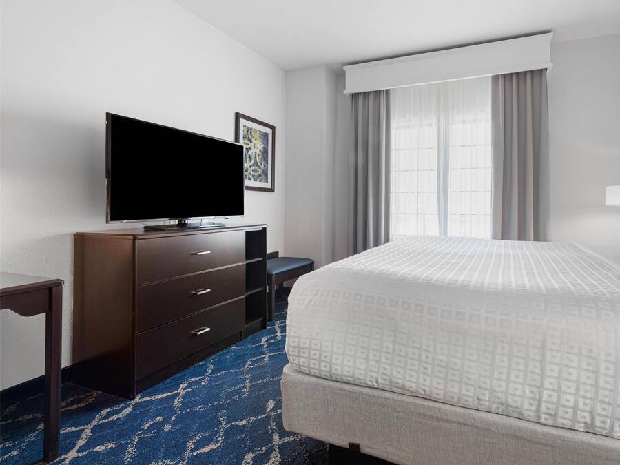 Habitación de hotel con una cama grande, un televisor de pantalla plana sobre un tocador de madera, un escritorio y una ventana con cortinas.