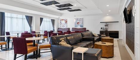 A modern hotel lobby with a black leather sofa, red chairs, and a white coffee table. There are multiple tables and chairs arranged for dining.