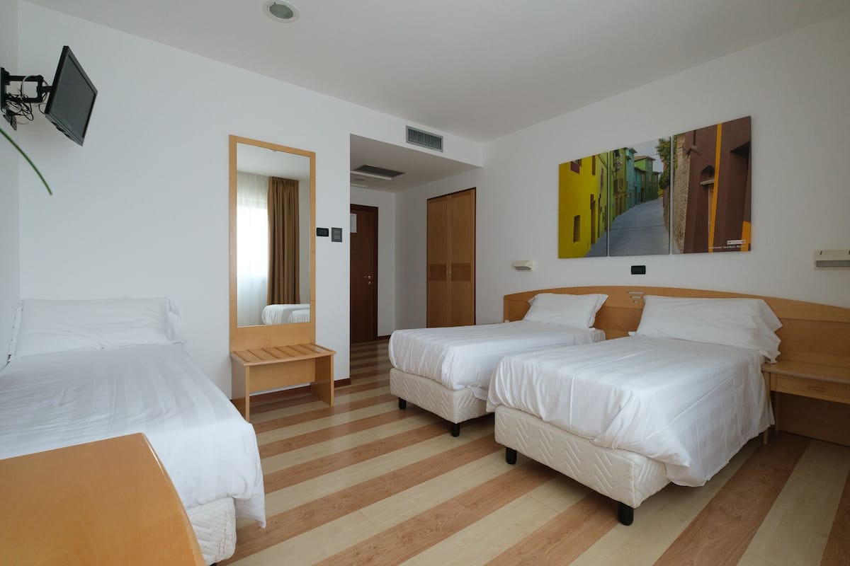 A hotel room with two single beds, a wooden bench, a TV mounted on the wall, and a painting above the beds.