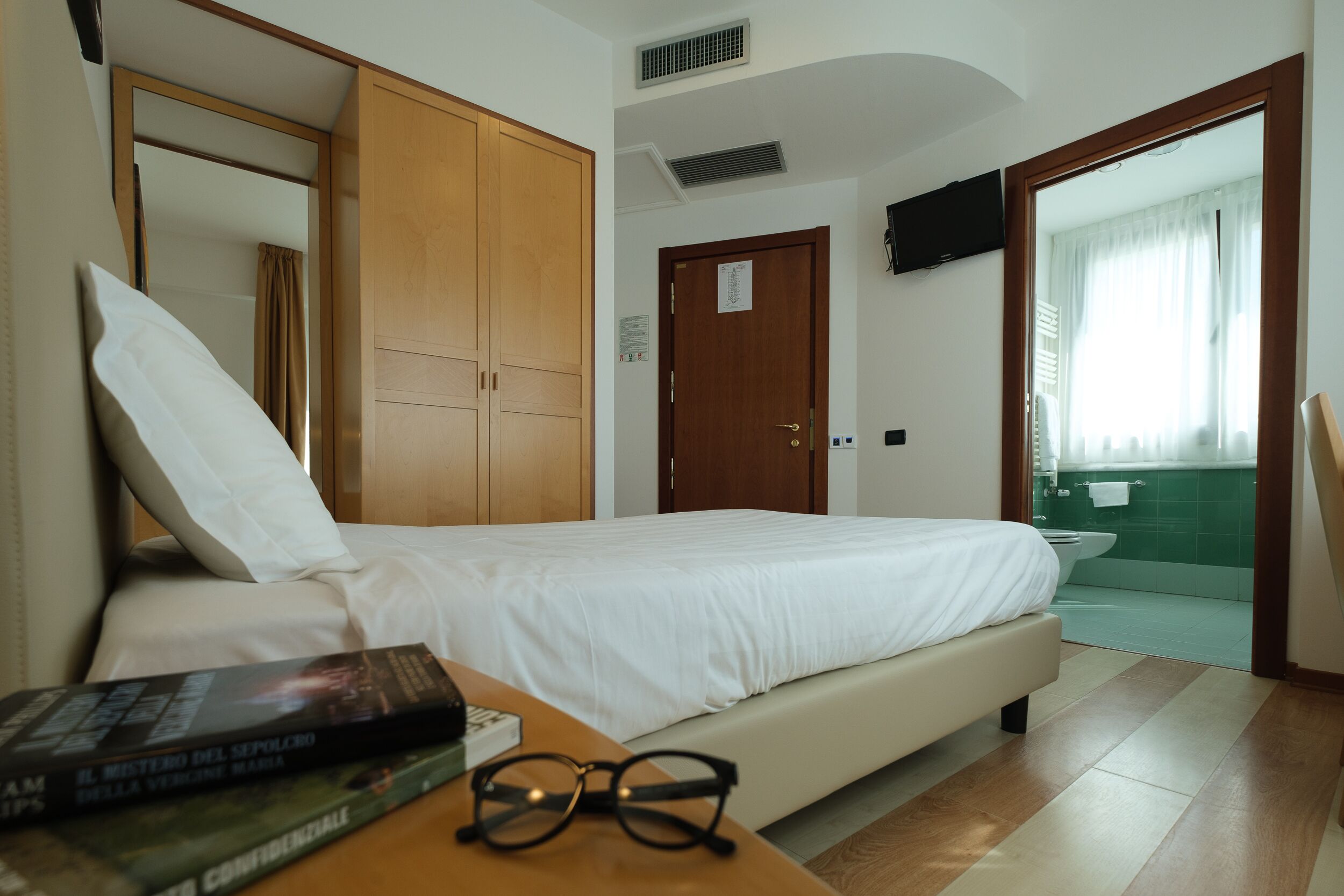 Habitación de hotel con cama, armario de madera, televisor montado en la pared y baño a la vista a través de una puerta abierta.
