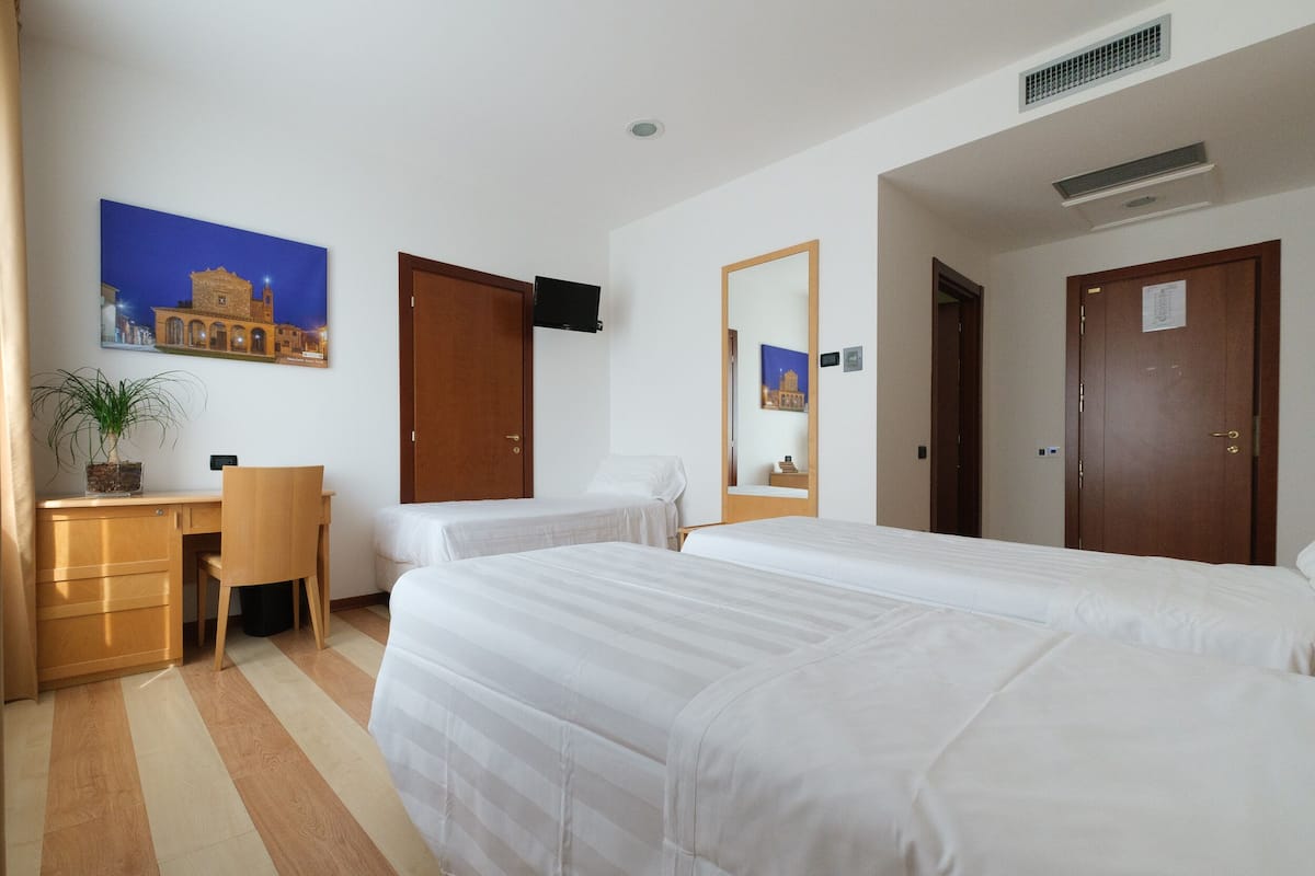 A hotel room with two beds, a desk, a chair, a potted plant, and a painting on the wall.