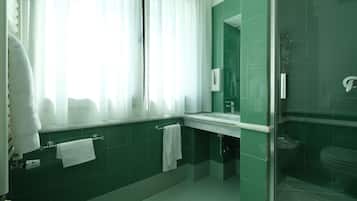 Un bany modern amb rajoles verdes, una dutxa amb envàs de vidre, un lavabo blanc i una finestra gran amb cortines transparents.