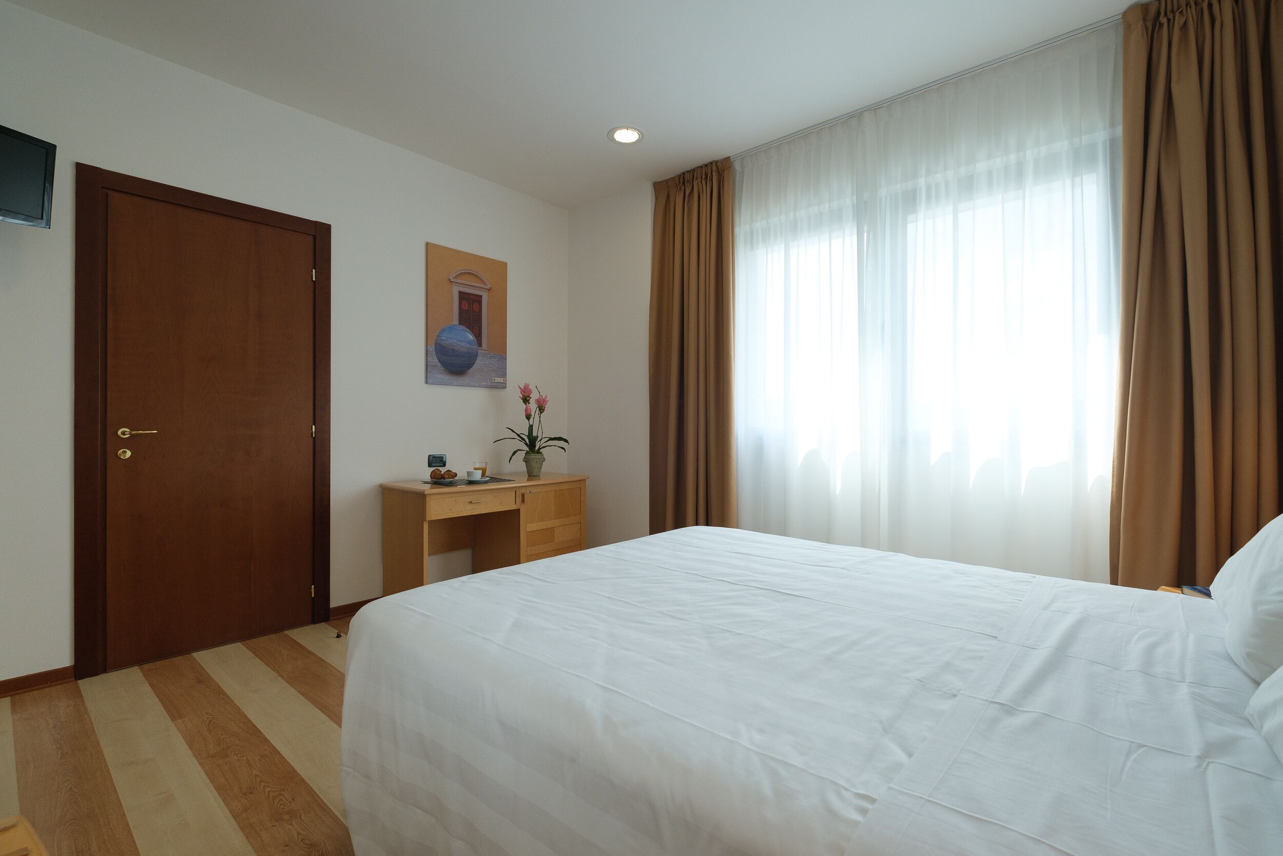 Une chambre d’hôtel avec un grand lit, un bureau en bois, une petite plante en pot et une porte.