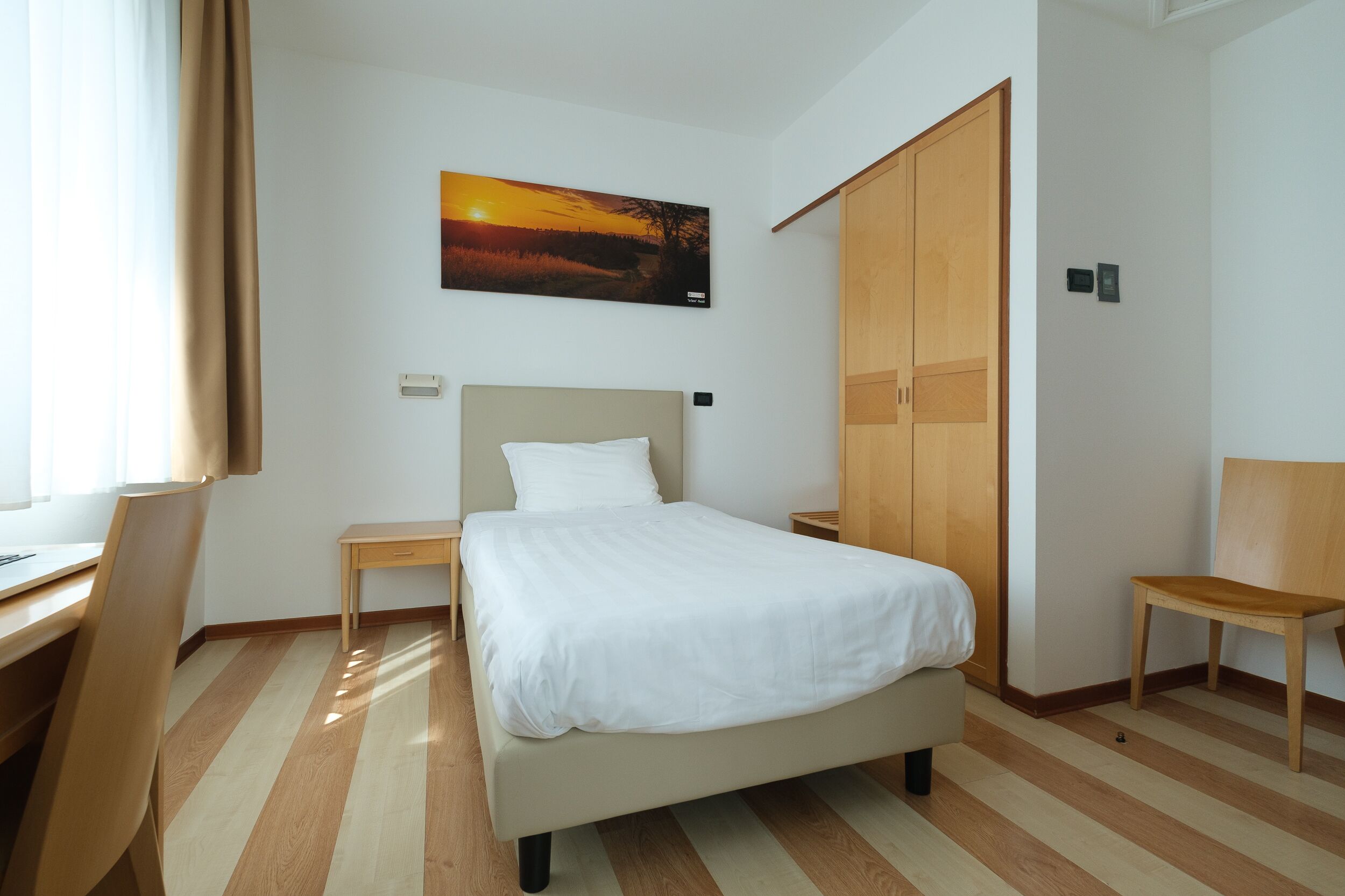A hotel room with a bed, a desk, a chair, a wardrobe, and a painting of a sunset.