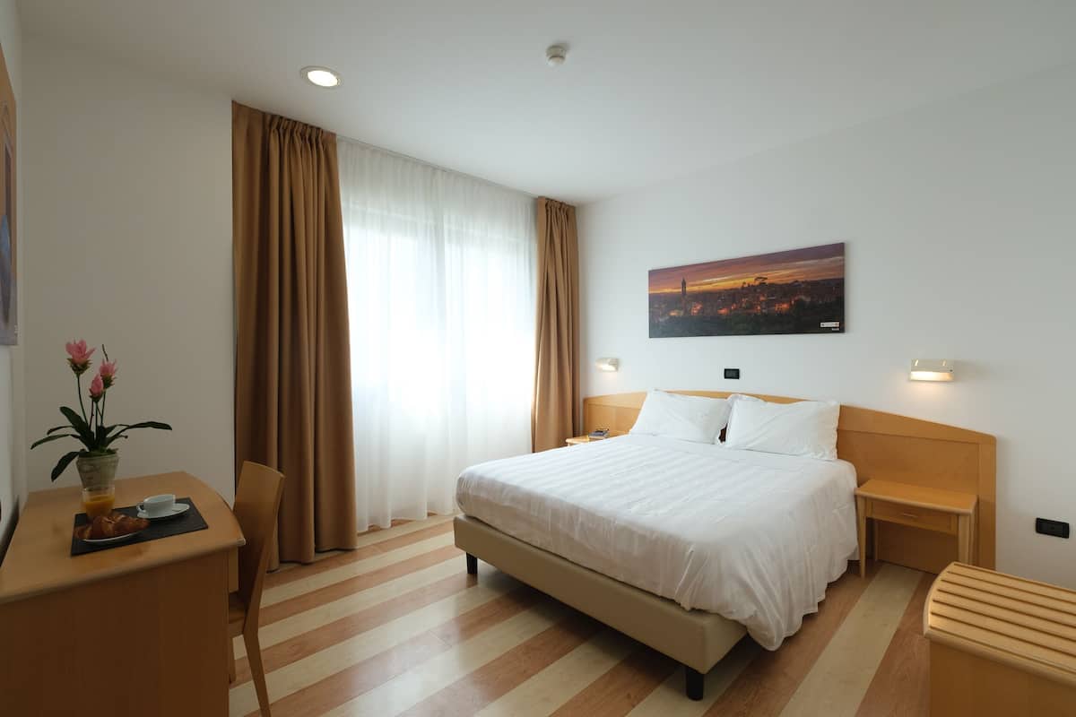 A hotel room with a large bed, a desk with a chair, a small table with a plant, and a painting on the wall.