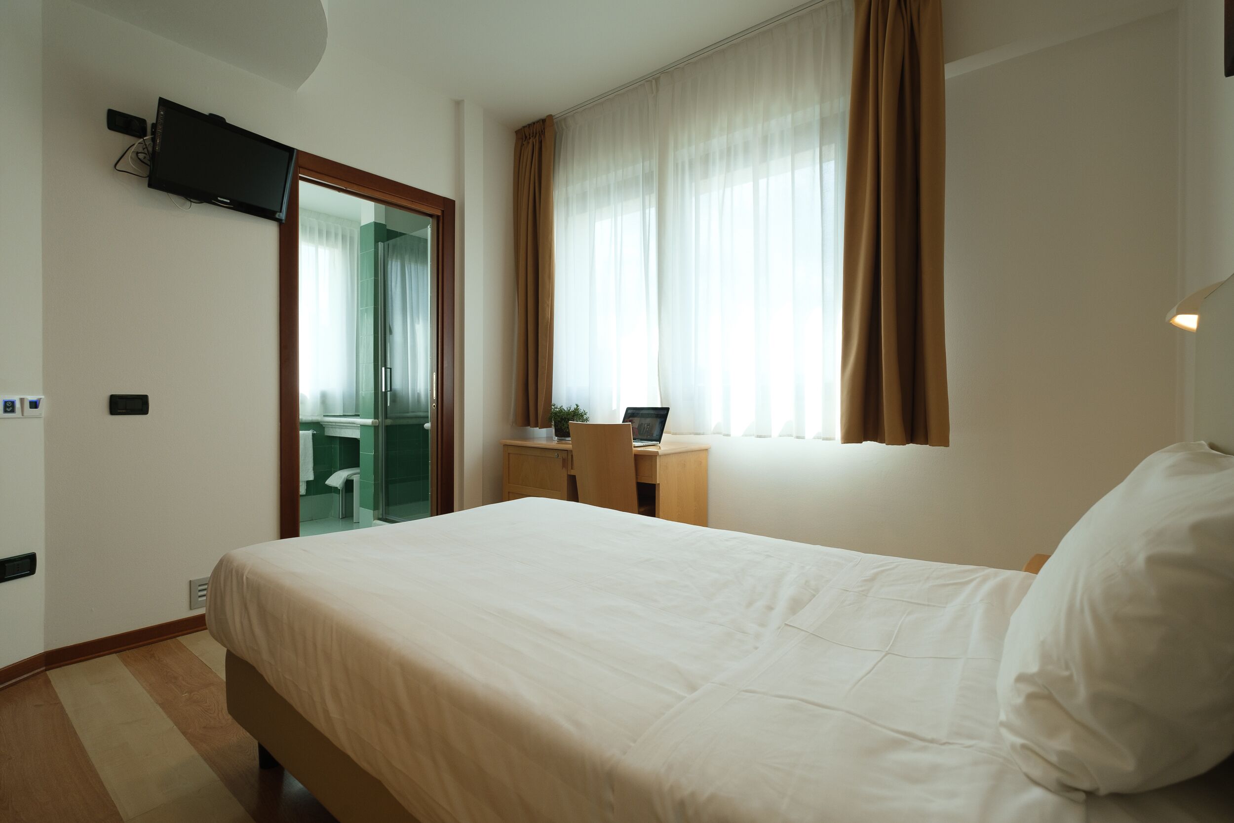 Habitación de hotel con una cama, un escritorio con una computadora portátil, un televisor montado en la pared y un baño visible a través de una puerta abierta.