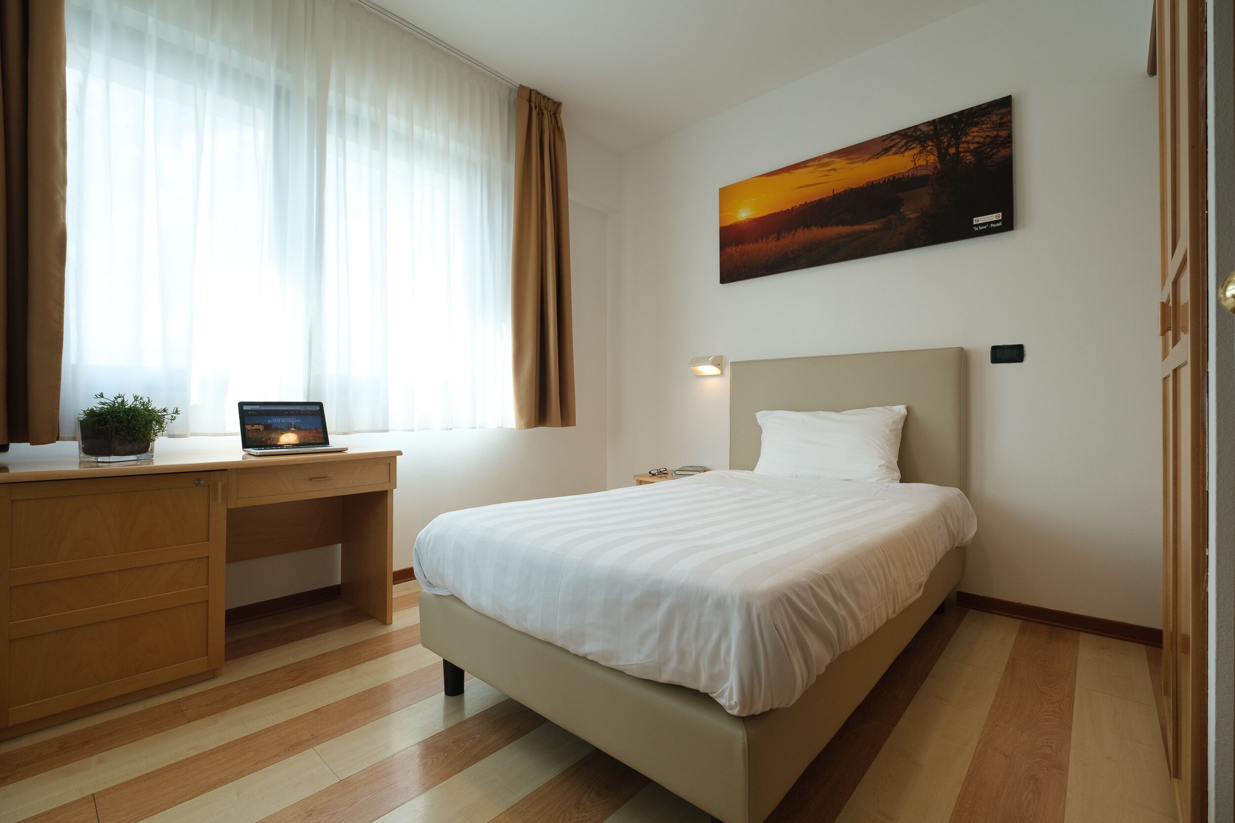Quarto com cama, escrivaninha, laptop e janela com cortinas.