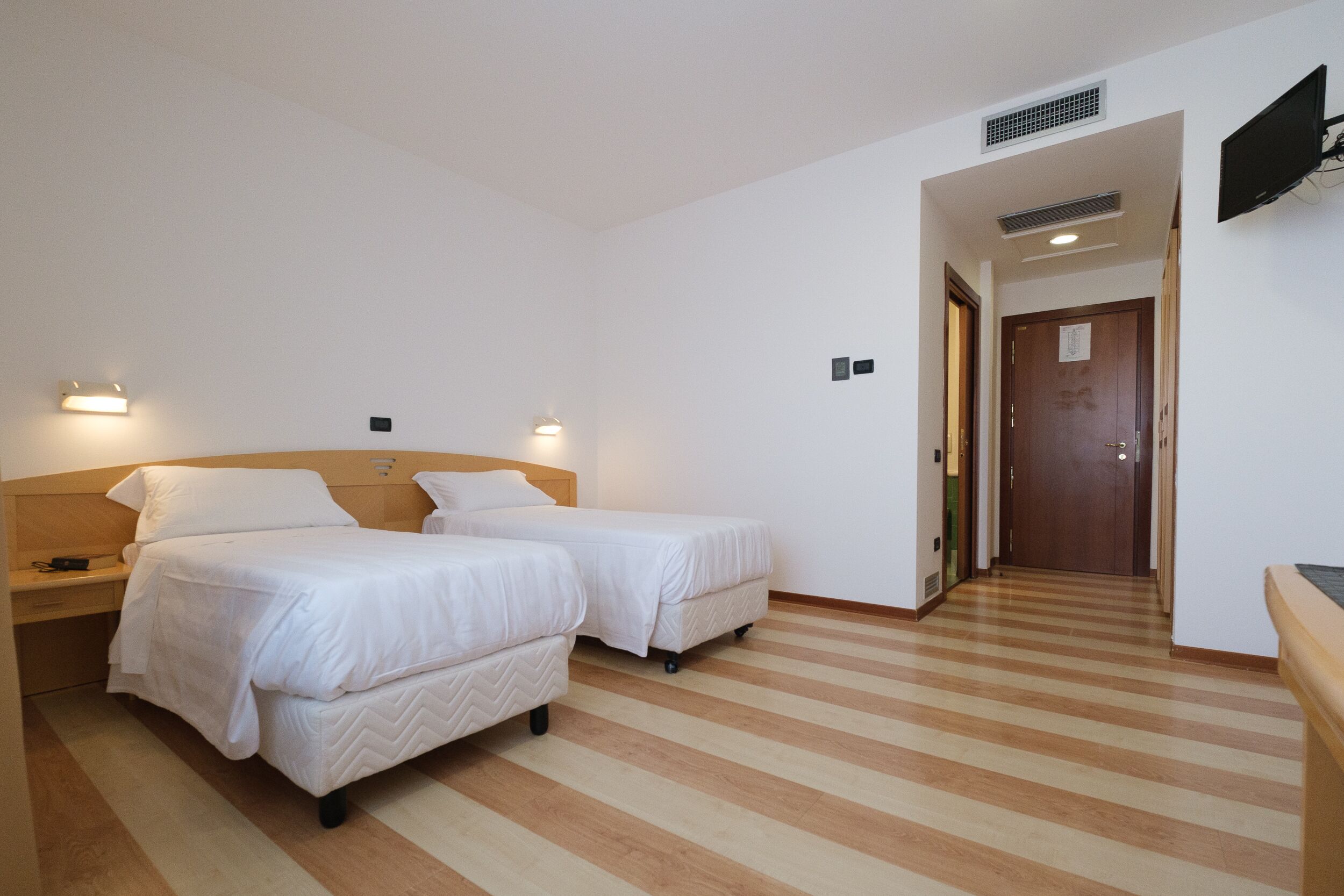A hotel room with two beds, a wooden floor, a TV mounted on the wall, and a door leading to another room.