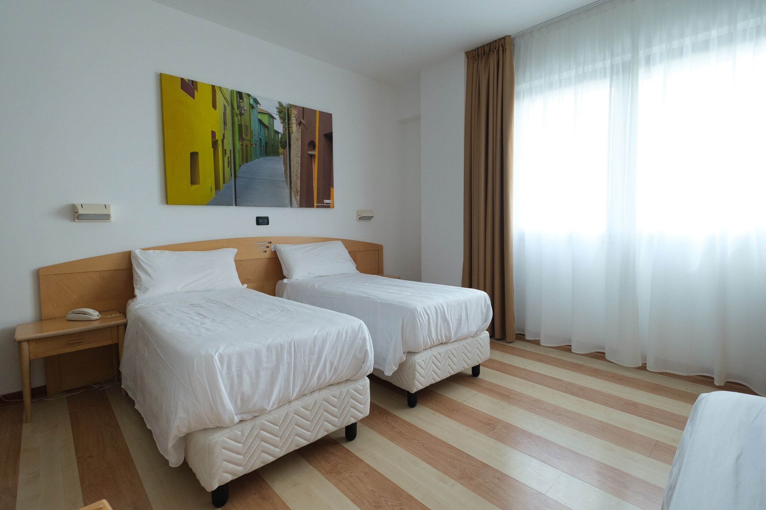Ein Hotelzimmer mit zwei Betten, einem hölzernen Kopfteil, einem Nachttisch mit Telefon, einem Gemälde an der Wand und einem großen Fenster mit Vorhängen.