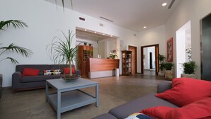 A modern living room with a grey sofa, a coffee table, and a red cushion. There is a wooden reception desk and a bookshelf in the background. - Hotel Portavaldera (Peccioli)