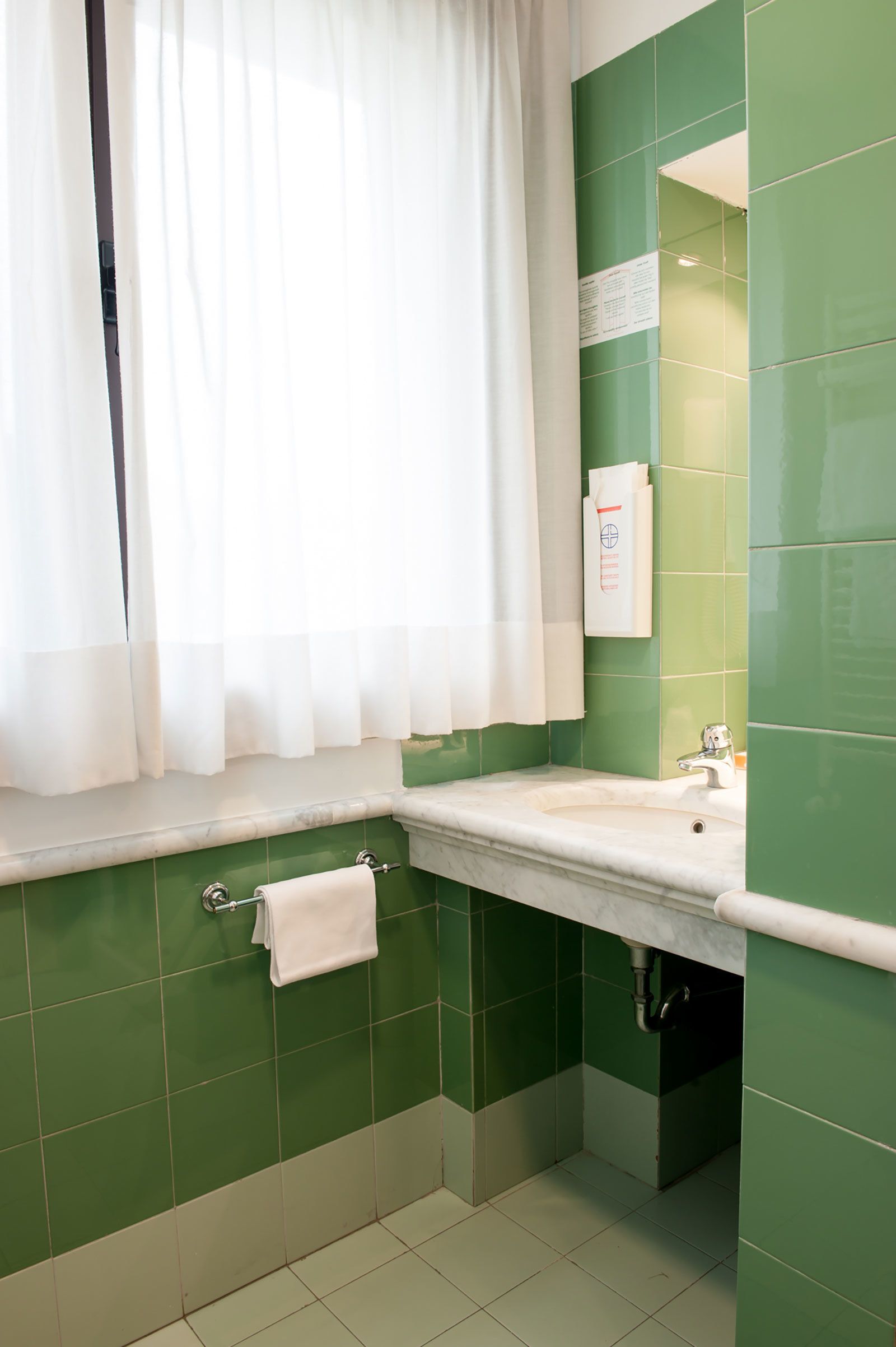 A bathroom with green tiles, a white sink, and a window with white curtains.