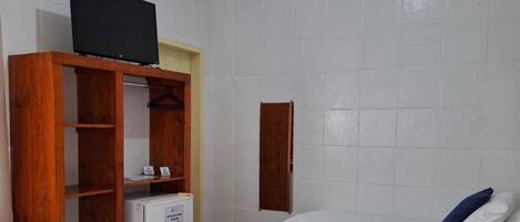 A hotel room with a bed, a TV on a wooden stand, a small refrigerator, and a white tiled wall.
