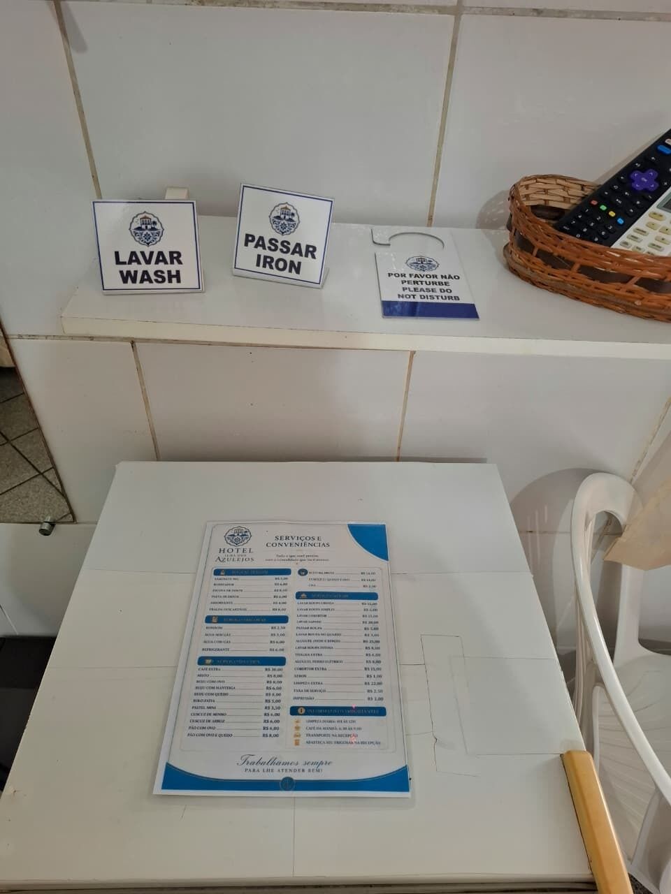 Een hotelkamer met een betegelde witte muur, een witte stoel, een tafel met een menukaart, en borden met 'LAVAR WASH' en 'PASSAR IRON'.