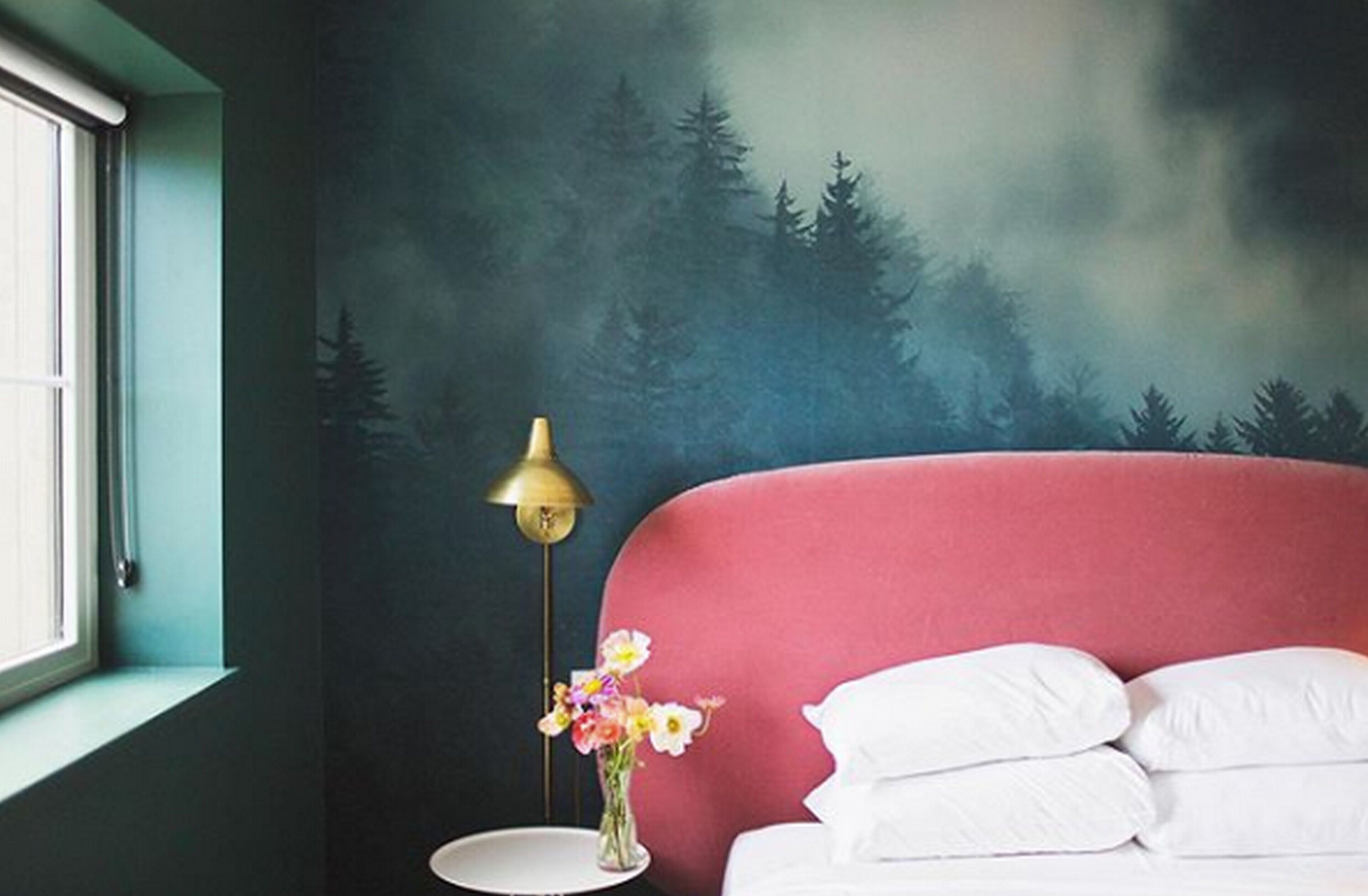 Un dormitorio con cabecera roja, almohadas blancas, una mesita de noche con lámpara y flores, y un mural en la pared con motivos forestales.