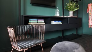 Un salon moderne avec un fauteuil en cuir noir, un pouf gris, un meuble de rangement noir contenant des livres et des objets décoratifs, et un téléviseur à écran plat fixé au mur.