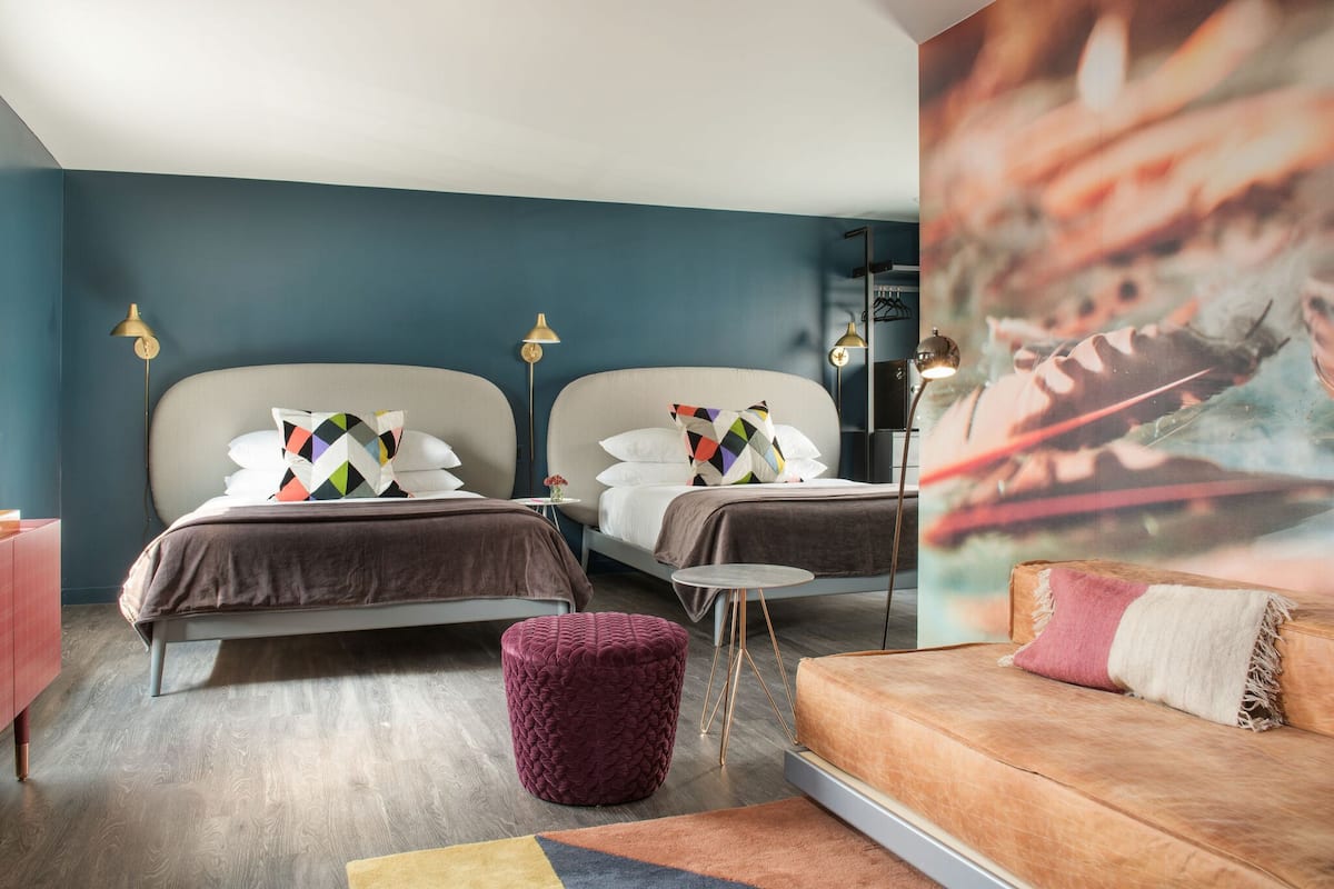 A modern bedroom with two beds, a small round ottoman, a sofa, and a painting on the wall.