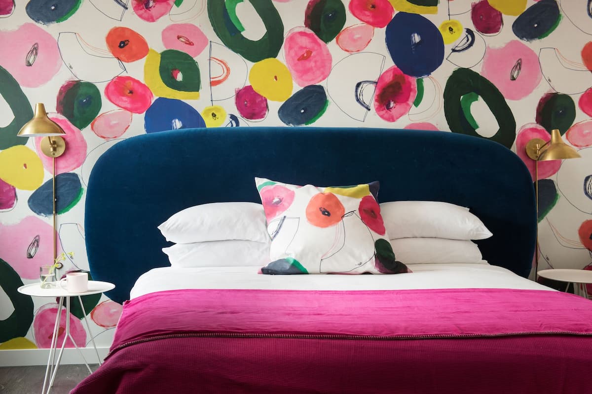 A bedroom with a vibrant floral wallpaper, a blue headboard, white and colorful pillows, a pink bedspread, and a small white table with a lamp.