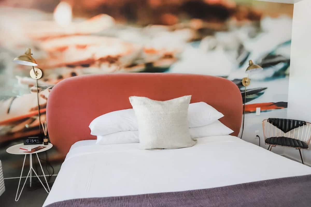 A hotel room with a bed, pillows, a small table with a clock, and a chair.