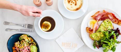 A breakfast setting with a variety of dishes, including toast with avocado and bacon, a cup of coffee with a lemon slice, and a bowl of salad.