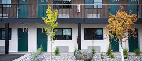 A building with numbered doors, likely a hotel or apartment complex, with a balcony and a gravel area with plants.