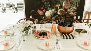 Une table à manger dressée avec des assiettes, des couverts, des verres et une variété d’aliments, notamment un plateau de fruits, des olives et une bouteille.