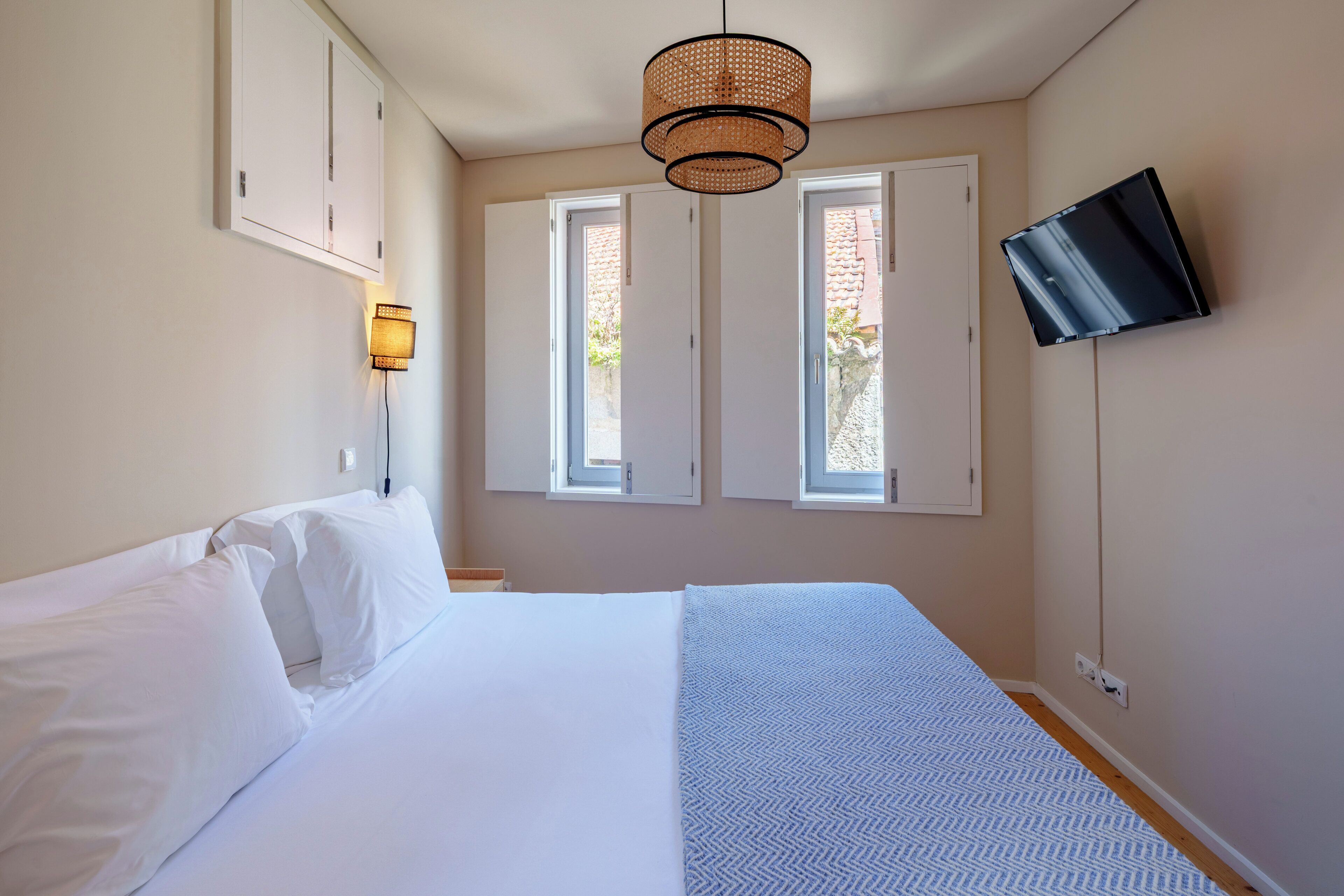Quarto com cama, duas janelas, TV fixada na parede e luminária pendente.