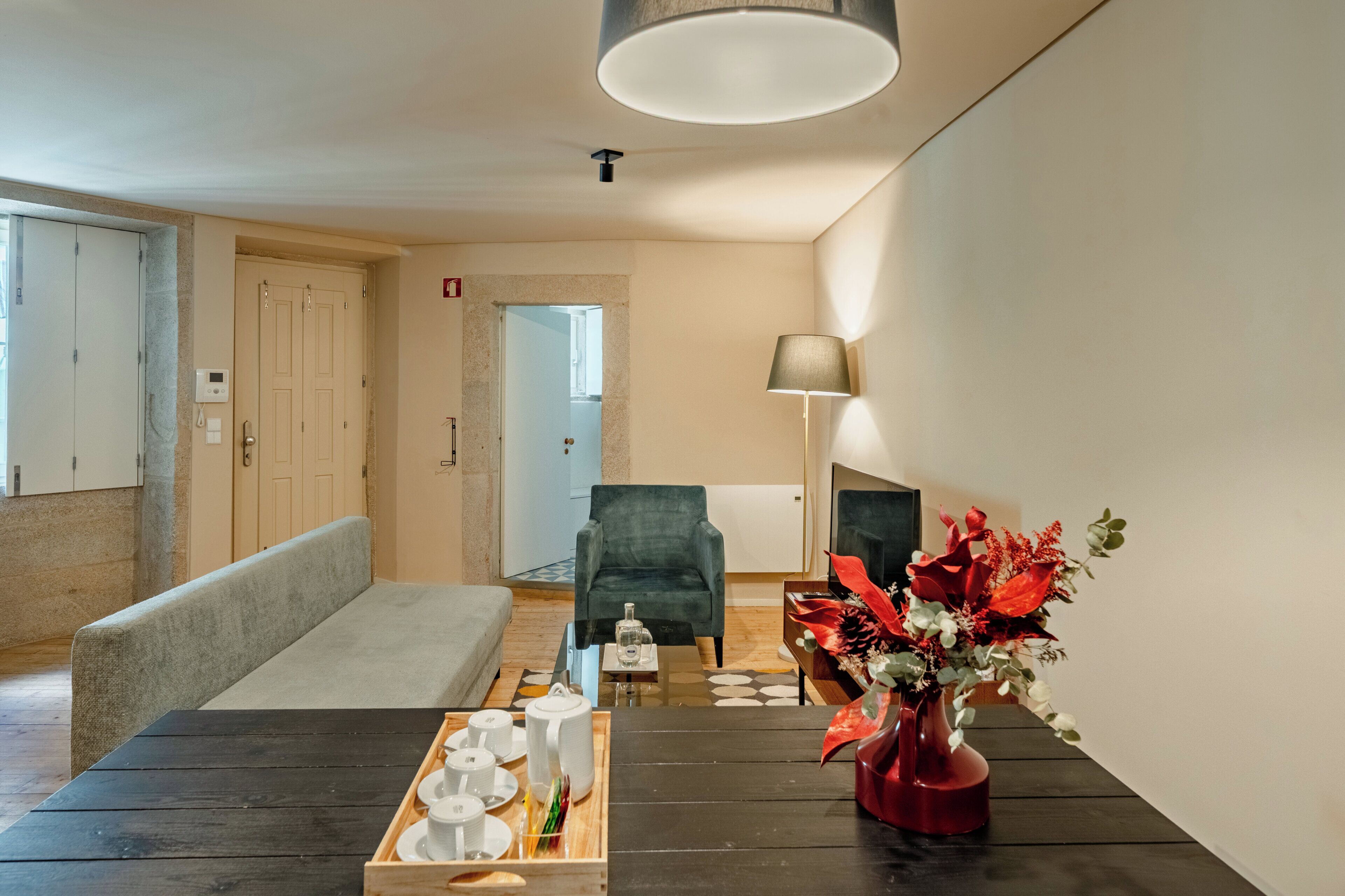 A modern hotel room with a wooden dining table, a bench, a sofa, a green armchair, a vase with flowers, and a lamp.