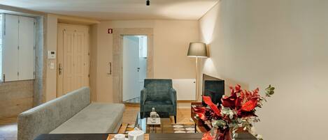 A modern hotel room with a wooden dining table, a bench, a sofa, a green armchair, a vase with flowers, and a lamp.