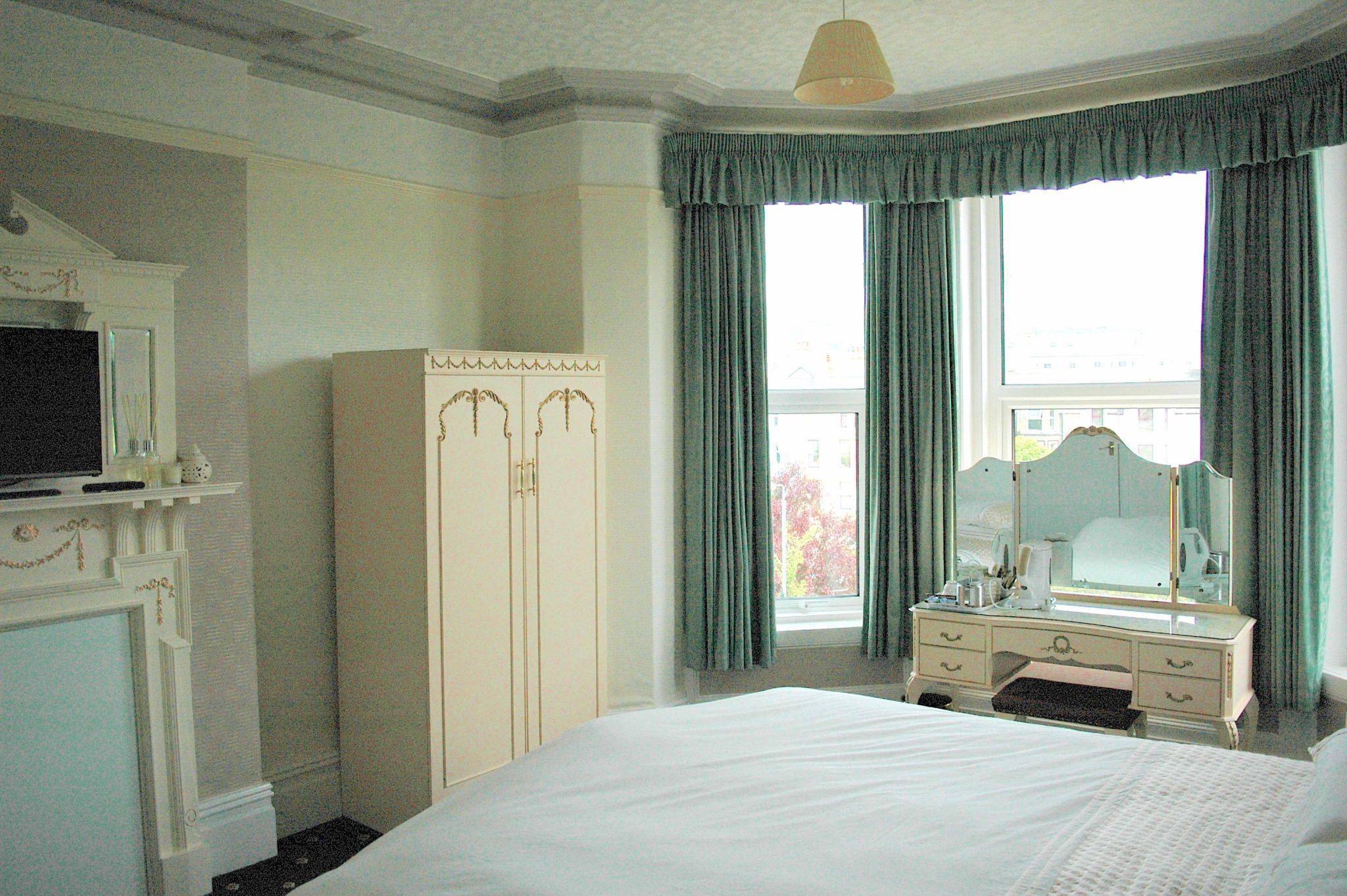 A hotel room with a large bed, a vanity with a mirror, a wardrobe, and a window with curtains.