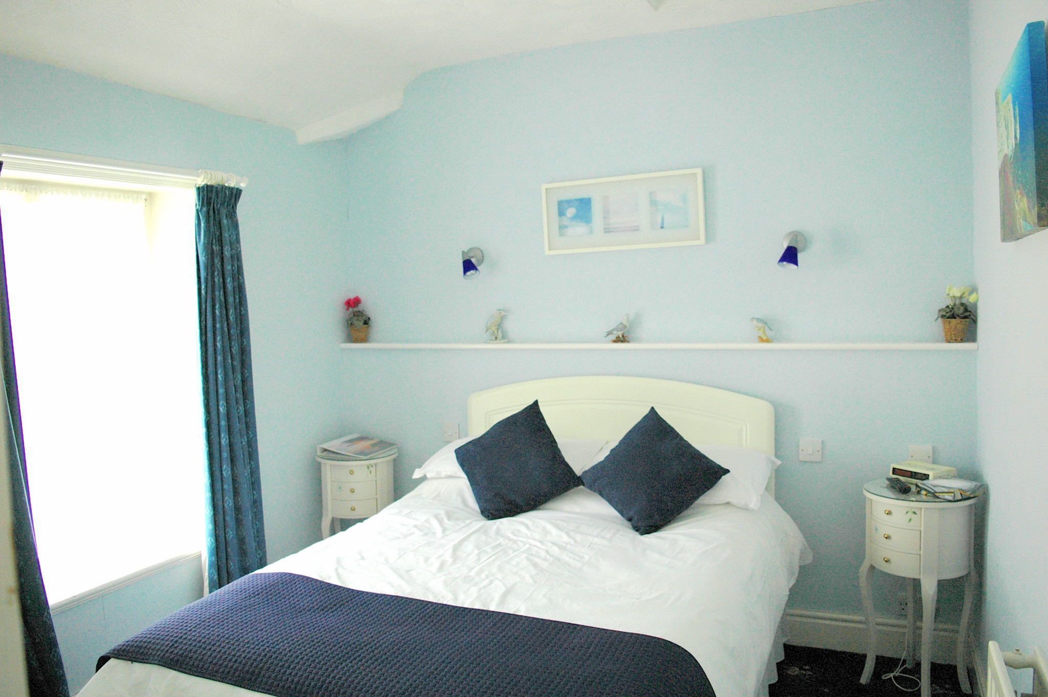 A bedroom with a bed, a nightstand, a shelf with decorative items, and a window with curtains.