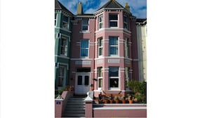 Un bâtiment de style victorien, peint en rose et en turquoise, doté d’une baie vitrée, d’une porte d’entrée et d’un petit porche agrémenté de plantes en pot.
