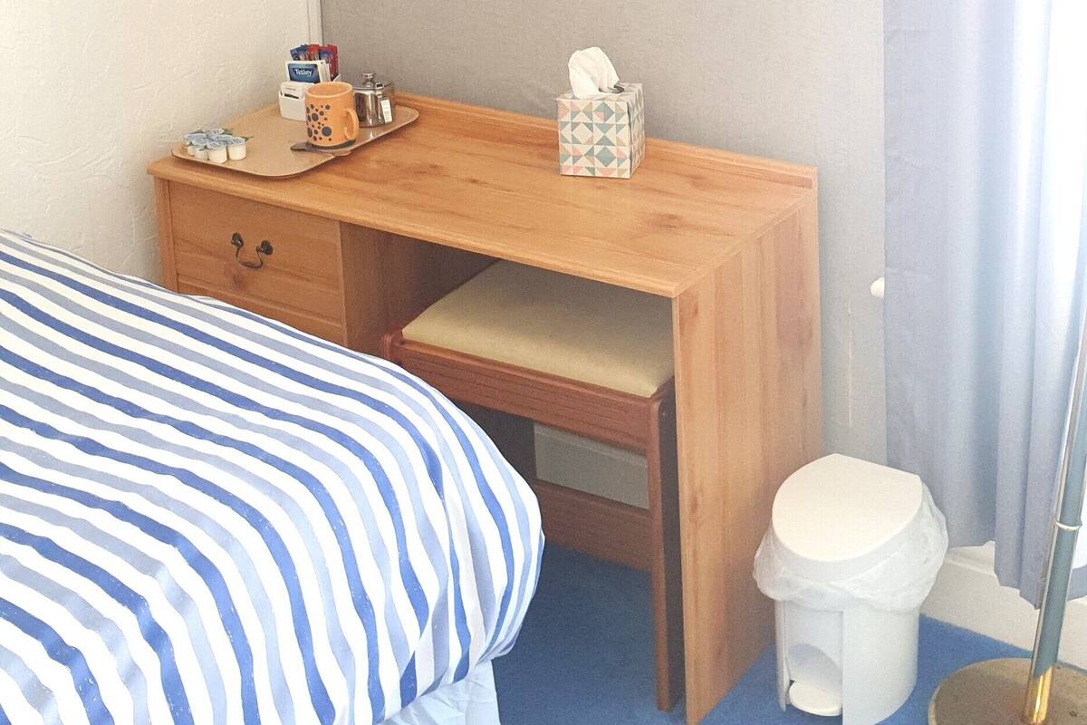 A hotel room with a bed, a desk with a flat-screen TV, a small stool, and a trash bin.