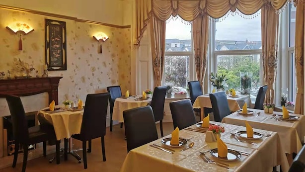 Un coin repas avec des tables dressées pour quatre personnes, chacune agrémentée d’une serviette jaune, de couverts et d’un vase de fleurs. On y trouve également deux lustres et une cheminée.