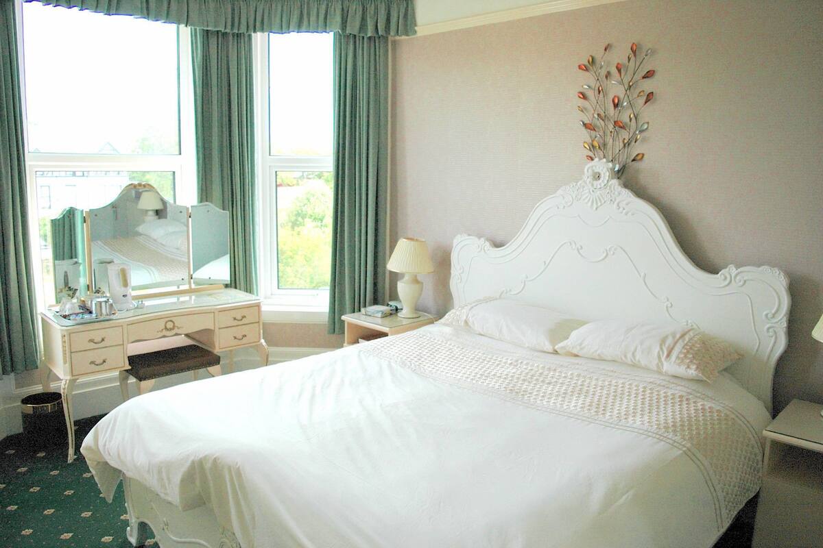 A neatly arranged bedroom with a large bed, a bedside table with a lamp, a dresser, and a window with curtains.