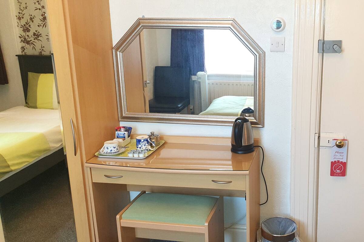 A compact hotel room with a single bed, a small desk with a mirror, a kettle, and a tray with tea and sugar.