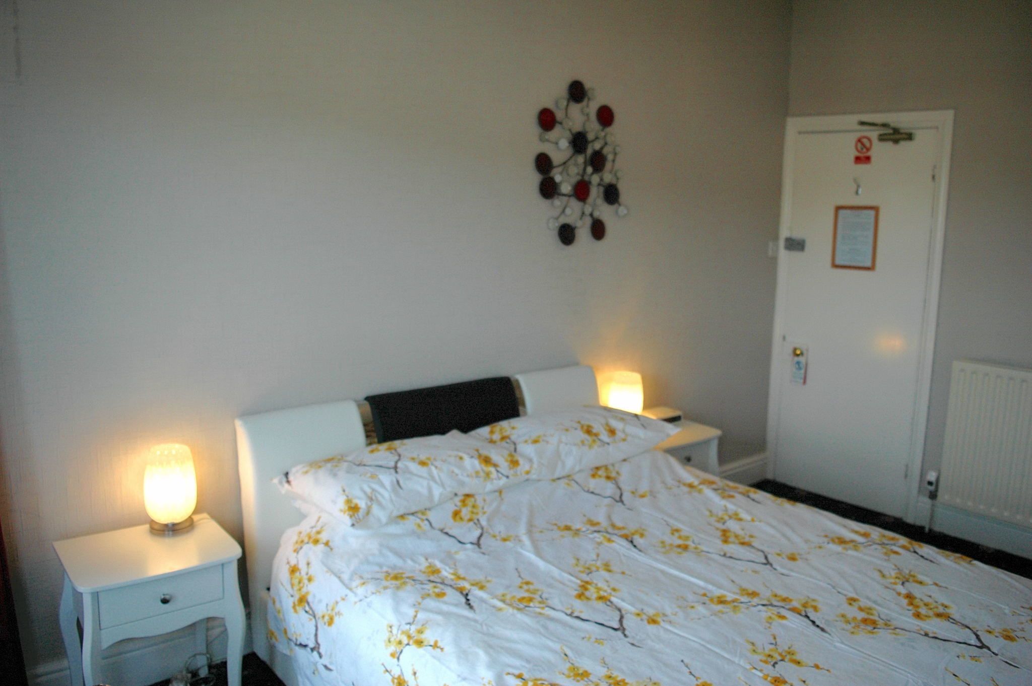 A bedroom with a bed, two bedside tables with lamps, a wall decoration, and a door with a sign.