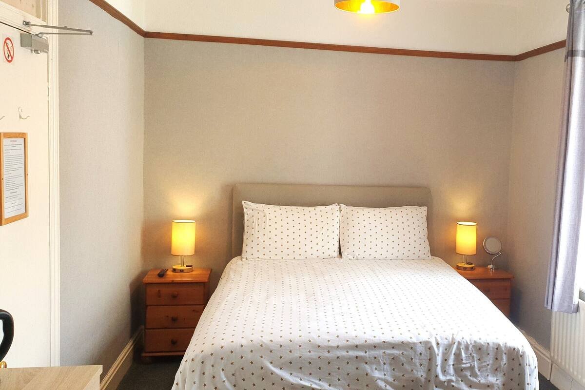 A bedroom with a bed, two bedside tables with lamps, a wooden headboard, a window with a curtain, and a wall-mounted light fixture.