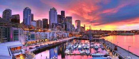 A marina with boats docked, a city skyline in the background, and a waterfront restaurant with outdoor seating.