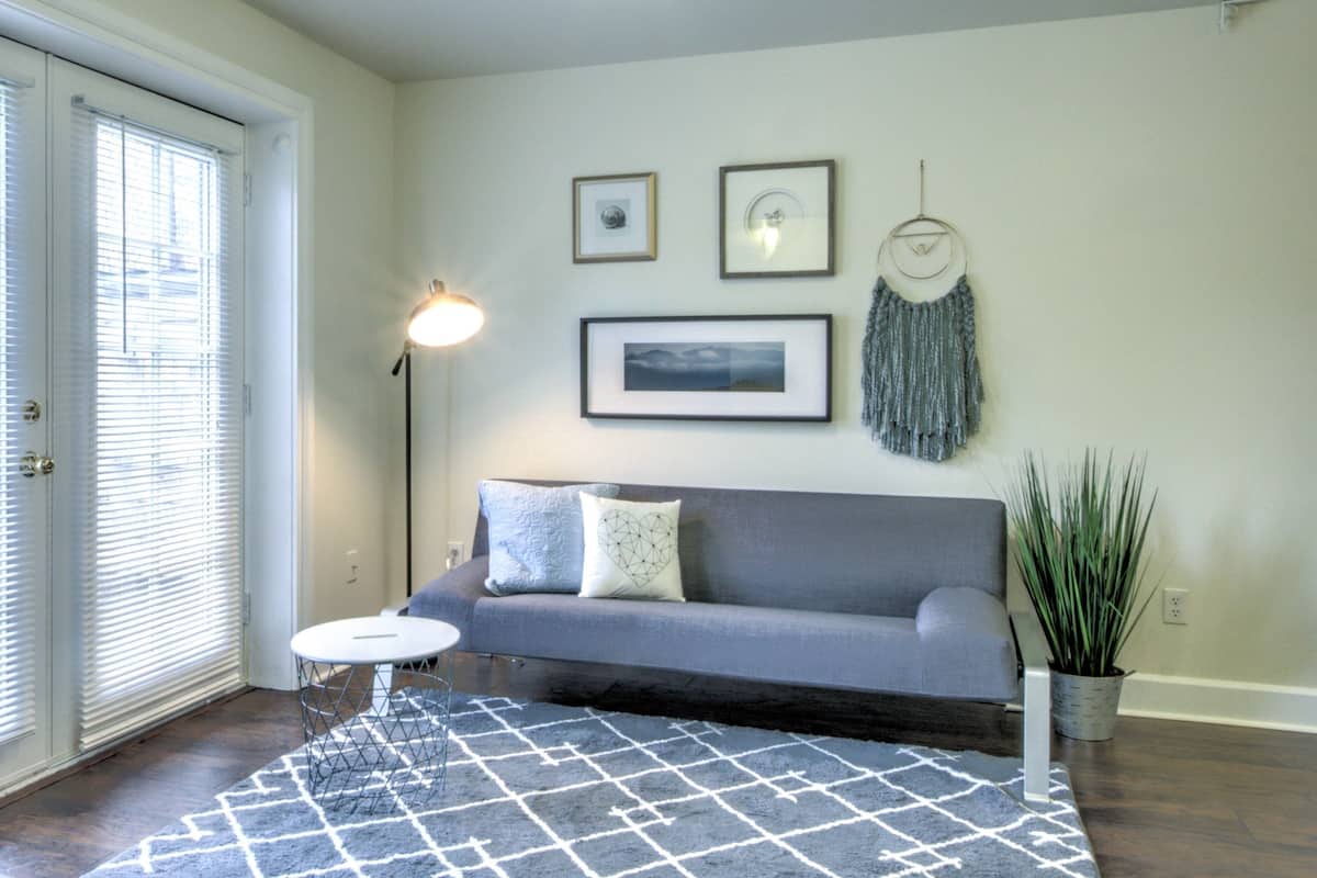 A living room with a gray sofa, a small round table, and a patterned rug. There are two framed pictures and a dreamcatcher on the wall.